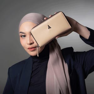 Puede incluir: Una cartera de cuero beige con ribete marrón oscuro y cremallera negra. La cartera tiene un logotipo negro en la parte delantera. La cartera se sostiene frente a la cara de una persona. La persona lleva una blazer azul marino y un velo beige.