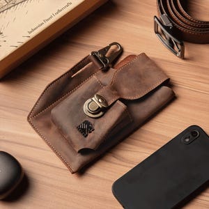 Könnte beinhalten: Eine braune Leder-Handyhülle mit Metallverschluss und Karabinerhaken. Die Hülle hat eine kleine Tasche und ein dekoratives Emblem. Ein schwarzes Smartphone liegt neben der Hülle. Der Text "Indonesian Finest Leathercrafts" ist sichtbar.