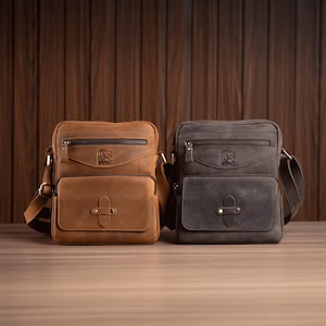 May include: Two leather messenger bags in brown and gray. Each bag has a front flap pocket, a zippered top, and a shoulder strap. The brown bag has a lighter tan color, while the gray bag is a darker shade. Both bags have a logo on the front.