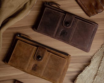 May include: Two leather wallets in shades of brown, one lighter and one darker, are displayed on a wooden surface. Each wallet features a zipper closure, a snap closure, and a logo. The wallets are rectangular and appear to be new.