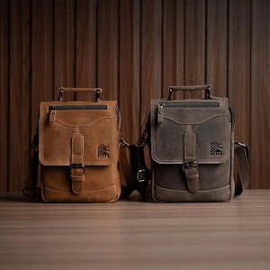 May include: Two leather messenger bags in shades of brown and gray. Each bag features a flap closure with a buckle, a top handle, and a shoulder strap. The bags have a textured finish and a logo on the front.