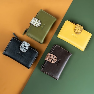May include: Four leather wallets in black, olive green, yellow, and brown are shown. Each wallet features a decorative clasp with a floral design and the word "alona" embossed on the front. The wallets are arranged on a two-tone background.