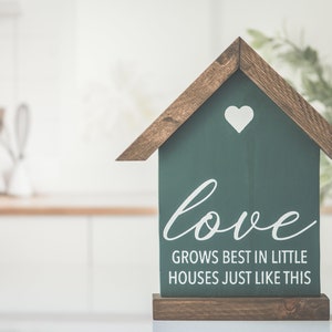 May include: A wooden house-shaped sign with a green background and white text that reads "love grows best in little houses just like this". A white heart is in the upper part of the sign.