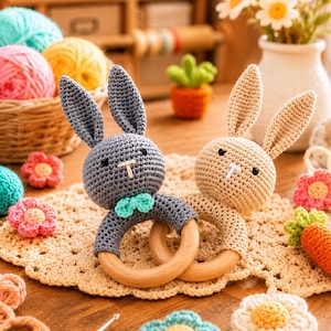 May include: Two crochet bunny rattles featuring gray and beige yarn heads attached to smooth, round wooden rings. The gray bunny has a small teal bow tie, while both have black embroidered eyes and white stitched noses, creating a pair of toys.