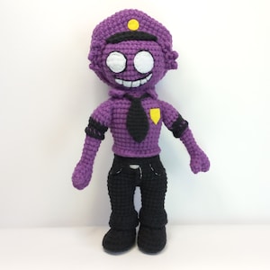 以下が含まれることがあります： 黄色いバッジ、黒いネクタイ、黒いズボンをつけた紫色の編みぐるみ。おもちゃは白い顔に黒い目と大きな笑顔をしています。