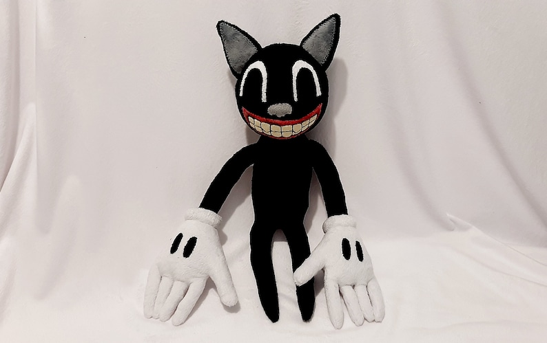 May include: A black cartoon cat plush toy with a wide, toothy grin. The cat has large white eyes and white gloves. The plush toy is sitting on a white surface.