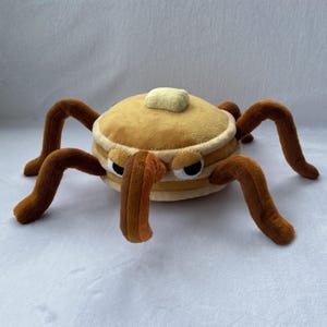 以下が含まれることがあります： 8本の茶色の脚が付いた、パンケーキの山を模したぬいぐるみ。パンケーキの山はタン色で、黄色いバターが乗っています。おもちゃは黒い目をしており、怒った表情をしています。
