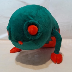Peut inclure: Un jouet en peluche en forme de créature verte et orange avec de grands yeux. Le jouet est couché sur le ventre, les bras tendus.