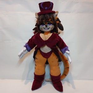 May include: A plush toy of a cat-like character wearing a purple and black hat, a red jacket, and white gloves. The character has orange fur, a purple nose, and a mischievous grin.