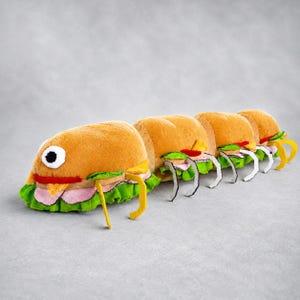 Könnte beinhalten: Ein skurriles Plüschtier in Form einer Raupe, bestehend aus fünf Sandwich-Abschnitten. Jeder Abschnitt hat ein Brötchen, Salat, Schinken und Käse. Die Raupe hat ein großes Auge und gelbe und weiße Beine.