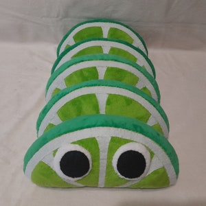 May include: A stack of five lime-shaped plush cushions with white accents and googly eyes. The cushions are arranged in a row, with the bottom cushion having the eyes.