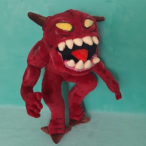 May include: Red plush toy demon with horns, yellow eyes, and a large open mouth with white teeth. The demon has a red tongue and is standing on two legs.
