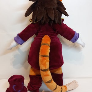 May include: A plush toy of a cat wearing a red velvet suit with a purple collar and a long orange and brown striped tail. The cat has brown hair and is standing on a white surface. A red velvet top hat with a purple symbol is on the surface.