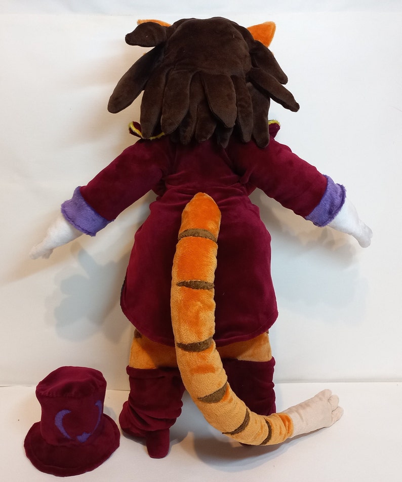 May include: A plush toy of a cat wearing a red velvet suit with a purple collar and a long orange and brown striped tail. The cat has brown hair and is standing on a white surface. A red velvet top hat with a purple symbol is on the surface.