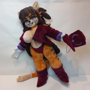May include: A plush toy cat with brown fur, a red and purple jacket, and a purple hat. The cat has a white chest, orange legs, and a long tail.