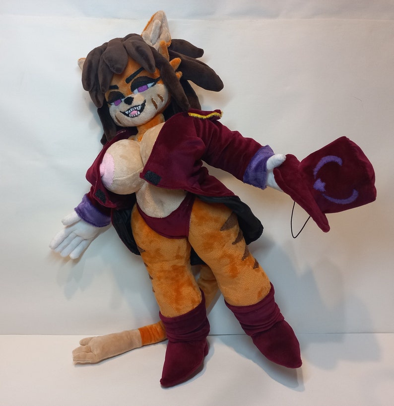 May include: A plush toy cat with brown fur, a red and purple jacket, and a purple hat. The cat has a white chest, orange legs, and a long tail.