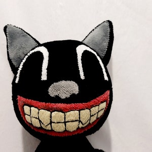 May include: A plush black cat toy with a wide, toothy grin. The cat has large, white eyes and a gray nose. The mouth is red with white teeth.