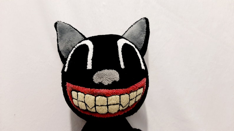 May include: A plush black cat toy with a wide, toothy grin. The cat has large, white eyes and a gray nose. The mouth is red with white teeth.