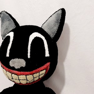 May include: A black plush cat with a large, toothy grin. The cat has white eyes and a gray nose. The cat's ears are gray and are sewn on.