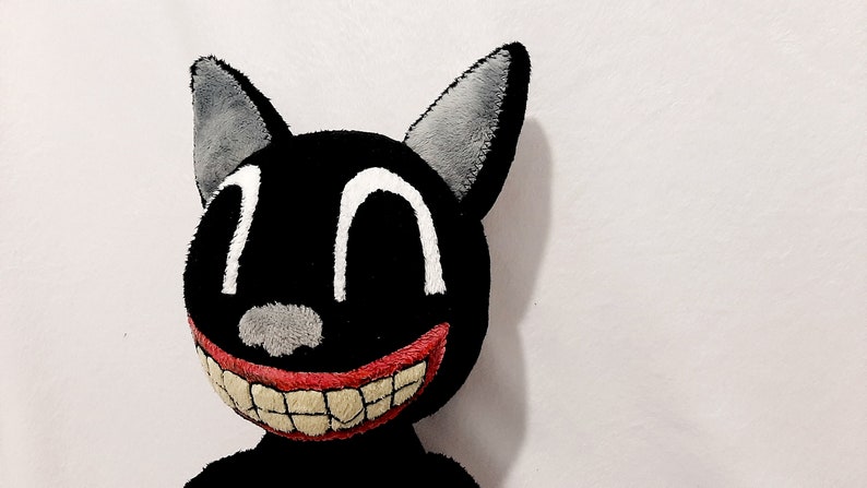 May include: A black plush cat with a large, toothy grin. The cat has white eyes and a gray nose. The cat's ears are gray and are sewn on.