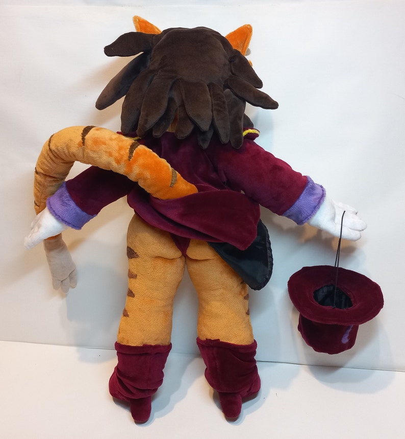 May include: A plush toy of a cat wearing a burgundy suit and holding a top hat. The cat has brown hair and a striped orange and brown tail.