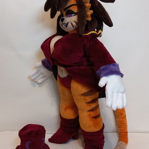 May include: A plush toy of a tiger-like creature with long brown hair, wearing a burgundy dress and white gloves. The toy has a long tail and is standing on a white surface. A burgundy hat with a purple heart design is in the foreground.