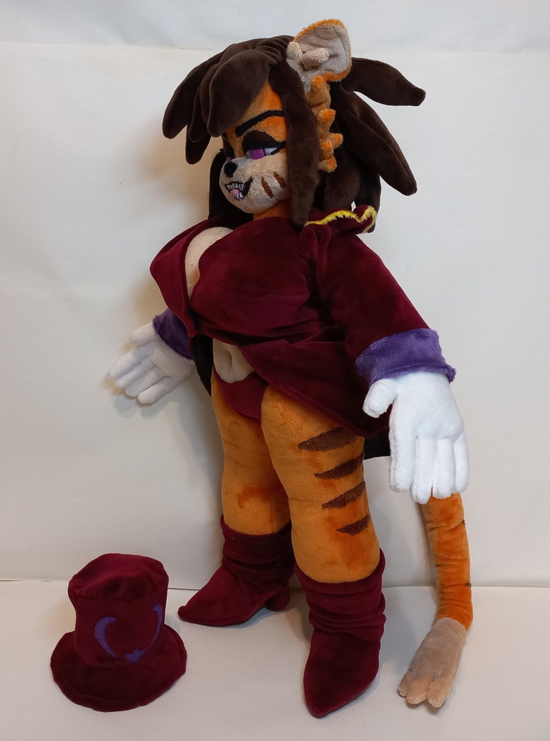 May include: A plush toy of a tiger-like creature with long brown hair, wearing a burgundy dress and white gloves. The toy has a long tail and is standing on a white surface. A burgundy hat with a purple heart design is in the foreground.