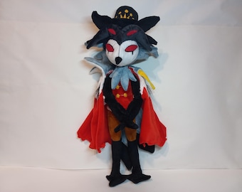 May include: A plush toy of a black and gray wolf wearing a red cape and a black crown. The wolf has red eyes and a white face. The toy is standing on a white background.