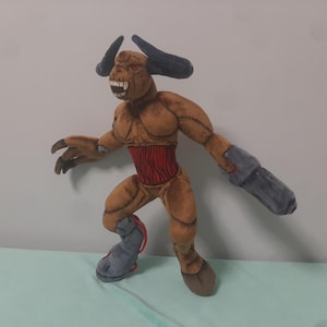 May include: A plush toy depicting a demonic creature with brown fur, black horns, and a red abdomen. The creature has a menacing expression with sharp teeth and a grey arm cannon. The toy is posed in a dynamic stance.