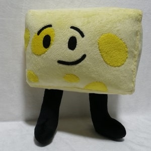May include: A plush toy shaped like a block of cheese with a smiling face and black legs. The cheese is yellow with brown spots and has a black smile and eyes.