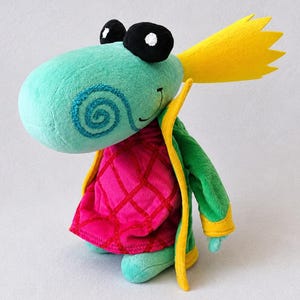 May include: A plush toy character with a light blue head, black eyes, and a yellow spiky crest. It features a blue spiral cheek detail, a green jacket with yellow trim, and a pink quilted vest with a red grid pattern over a green body.