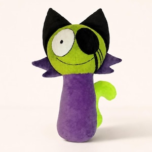 Puede incluir: Un juguete de peluche con forma de gato con una cara verde lima, orejas negras y un cuerpo morado. El gato tiene un ojo cubierto con un parche negro y una sonrisa cosida. La cola es verde lima.