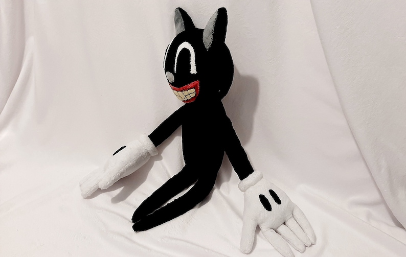 May include: A black plush cartoon cat with a large, toothy grin. The cat has white gloves and is sitting on a white surface.