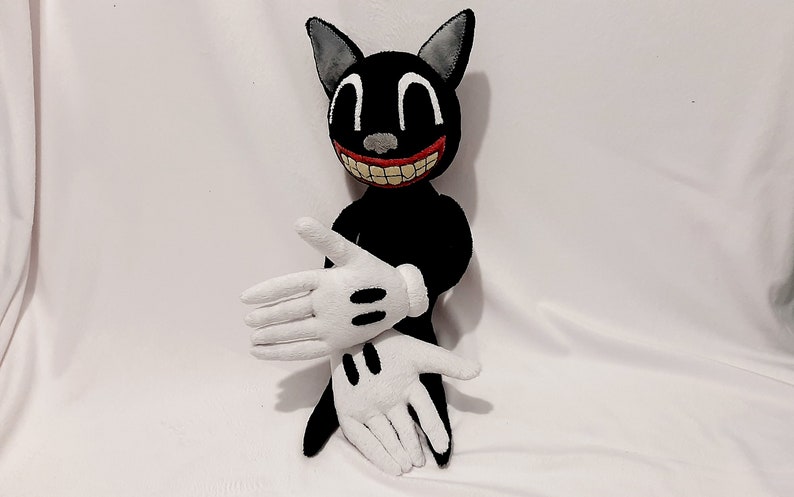 May include: A black cartoon cat plush toy with a wide, toothy grin. The toy has white gloves and is sitting on a white surface.