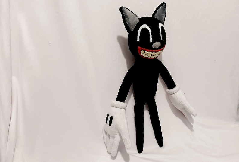 May include: A black cartoon cat plush toy with a wide, toothy grin. The toy has white gloves and large, white eyes. The toy is standing on a white surface.