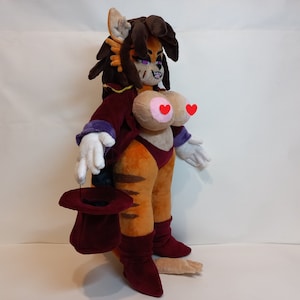 May include: A plush toy of a female character with orange fur, brown hair, and a red and purple outfit. She is holding a red top hat in her hand.
