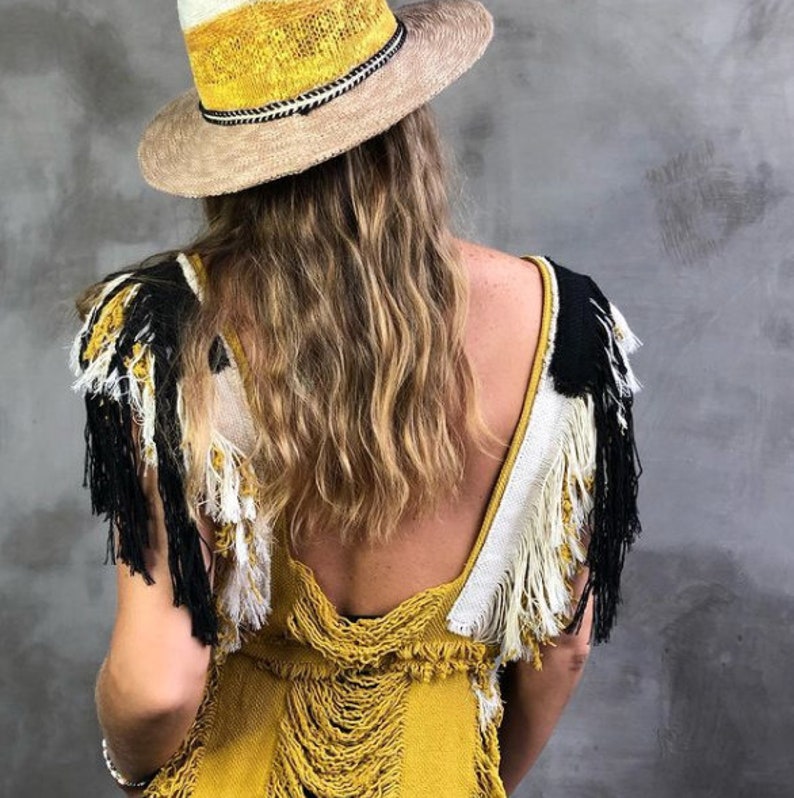 Op de afbeelding: Een vrouw draagt een gele gehaakte jurk met zwarte en witte franjes. Ze draagt een brede strohoed met een gele en witte band.