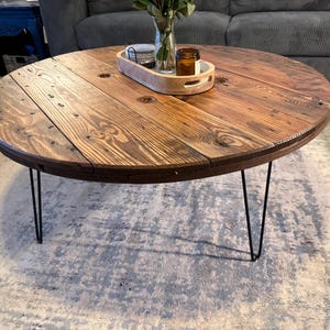 May include: Round wooden coffee table with a dark stained finish. The tabletop is made of planks and has a natural wood grain. The table has black hairpin legs. A wooden tray with decorative items sits on the table.