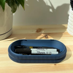 May include: An oval-shaped, dark blue desk organizer tray with a ribbed, textured exterior. The tray contains two white Sharpie Ultra Fine Point permanent markers and a black pen, resting horizontally inside the basin of the container.