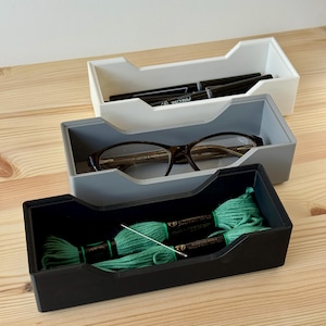 May include: Three rectangular storage trays in white, gray, and black, stacked on a wooden surface. The top tray holds black pens, the middle tray contains eyeglasses, and the bottom tray has green embroidery floss and a needle. A practical storage solution.