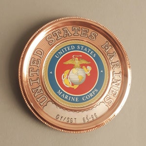 May include: A copper-colored decorative plate featuring the United States Marine Corps emblem. The emblem is centered on a blue and red background, surrounded by the words "United States Marines". The plate also includes the text "GY/SGT 65-86".