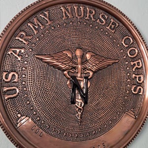 May include: A copper-colored, circular emblem with the text "US ARMY NURSE CORPS" around the edge. The center features a medical symbol with wings and a snake. The bottom of the emblem reads "2000-2025".