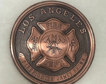 Può includere: Una moneta rotonda di colore rame con un emblema del Dipartimento dei Vigili del Fuoco di Los Angeles. L'emblema presenta una croce rossa con un'ascia, una scala e un idrante. Il testo "Los Angeles Fire Dept. Firefighter James Brown" è inciso attorno all'emblema. La moneta ha un diametro di 35 cm.