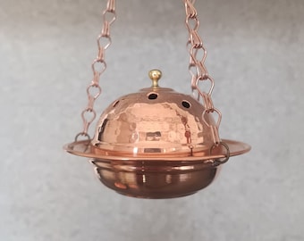 Op de afbeelding: Een koperen hangende wierookbrander met een gehamerde afwerking en een ketting om op te hangen. De brander heeft een ronde vorm met kleine gaatjes voor wierook om doorheen te branden.