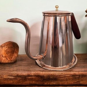 May include: A silver stainless steel pour-over coffee kettle with a wooden handle on a rustic wooden shelf.