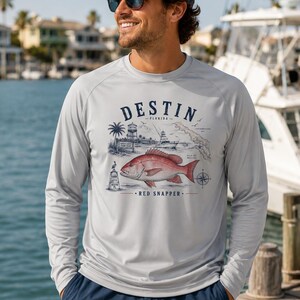 Puede incluir: Una camiseta de manga larga gris claro con un estampado gráfico de un pargo rojo, un barco, una palmera y un faro. El texto dice DESTIN FLORIDA y RED SNAPPER. La camiseta tiene cuello redondo y mangas raglán, ofreciendo un estilo casual.