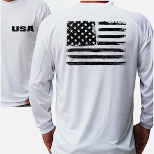 May include: A white long-sleeve shirt featuring a black distressed American flag graphic on the back and the text USA printed in black on the front left chest. The shirt has a crew neckline and long sleeves with a smooth, lightweight fabric finish.