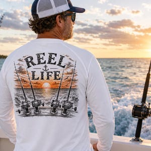 May include: A white long-sleeve shirt featuring a graphic on the back with the text "REEL LIFE" and an anchor icon. The illustration depicts four fishing rods positioned over a sunset ocean scene with waves and birds in a black and orange ink style.