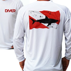 Puede incluir: Camiseta blanca de manga larga con texto "DIVER" rojo en la parte delantera. La parte trasera presenta un diseño de bandera roja y blanca desgastada con una silueta de tiburón negro. La camiseta es de material ligero y transpirable.