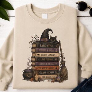May include: A beige crewneck sweatshirt featuring a graphic print of a stack of books titled Basic Witch, Potions & Spells, Book of Shadows, Love Potions, Curses & Hexes, The Witches Guide, Tarot Secrets, and Herbology, with a black cat and hat.
